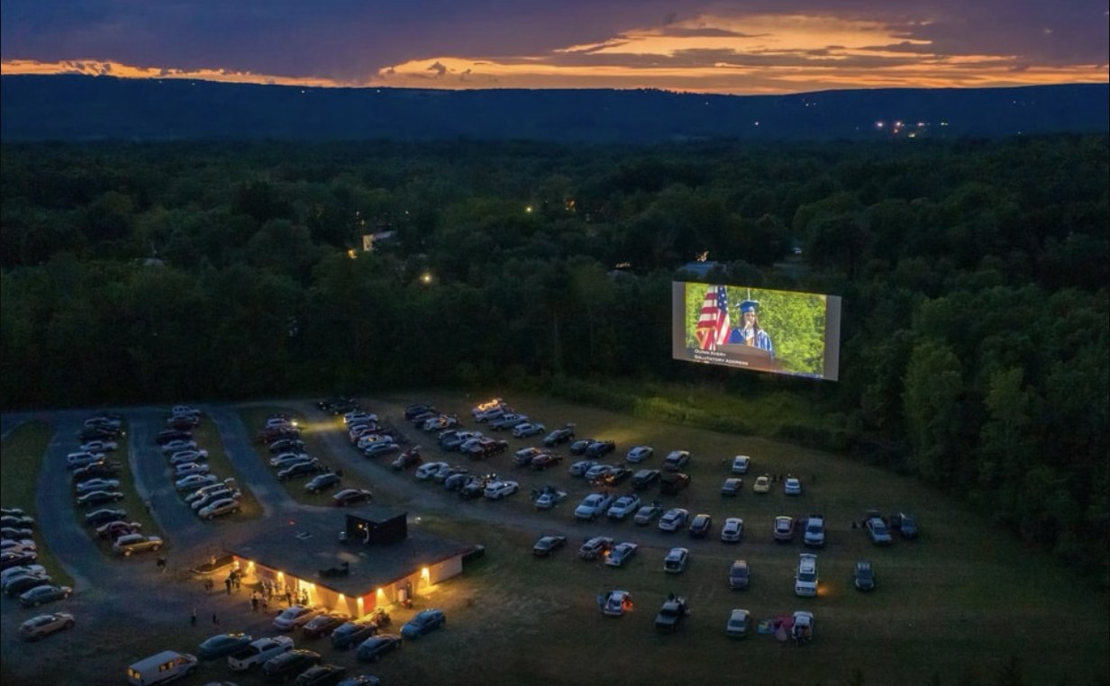 Aerial view of the Fair Oaks Drive-In at dusk, cars parked in rows facing a lit screen under an orange sunset.