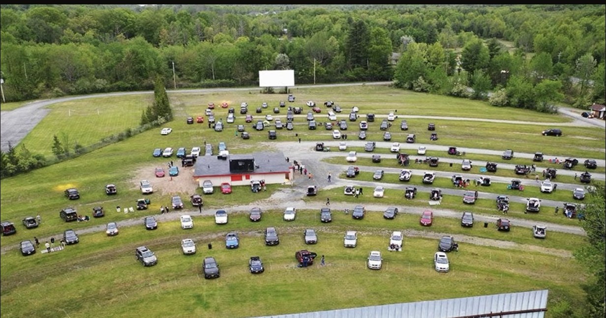 Daytime aerial view of the drive-in field with cars parked across the grass and the second screen in the foreground.